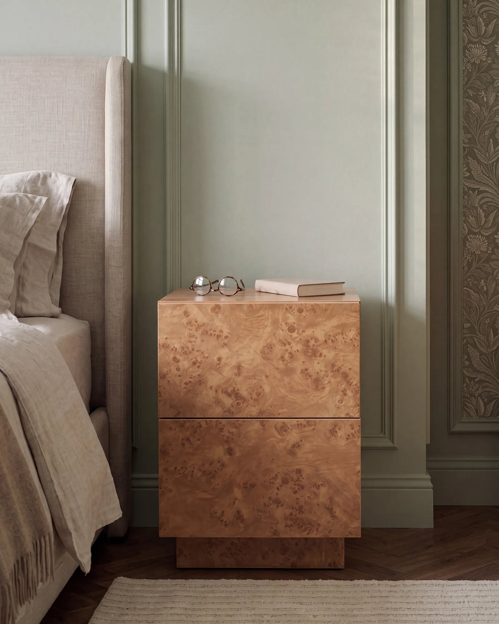 A two-drawer nightstand with a rich burl wood grain finish in warm brown and gold tones, styled in a bedroom next to a bed with neutral beige bedding.