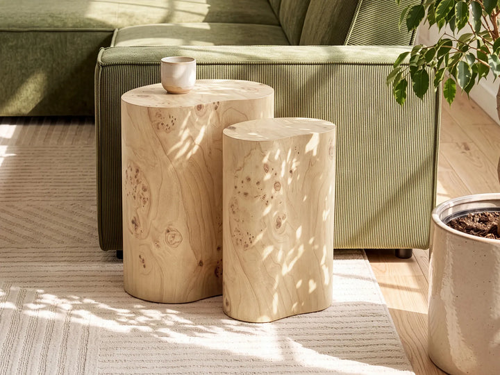 Two natural burl wood cylindrical side tables in a modern living room setting, with one smaller table in front of a larger one, set on a striped rug in front of a green sofa.