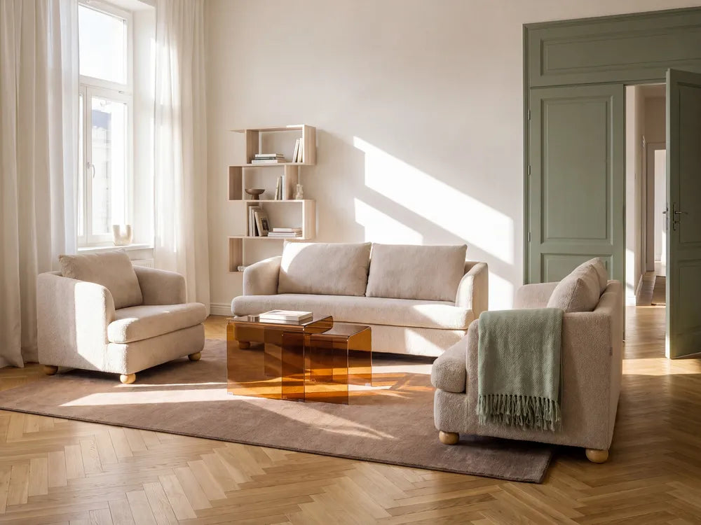 A styled living room featuring a light beige sofa, a matching armchair, a second accent chair with a green throw blanket, a translucent amber coffee table, and a brown rug on a herringbone wood floor.