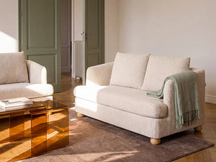 A cream-colored textured bouclé loveseat sofa in a bright, modern living room with warm sunlight, a green throw blanket, and a translucent amber glass coffee table.