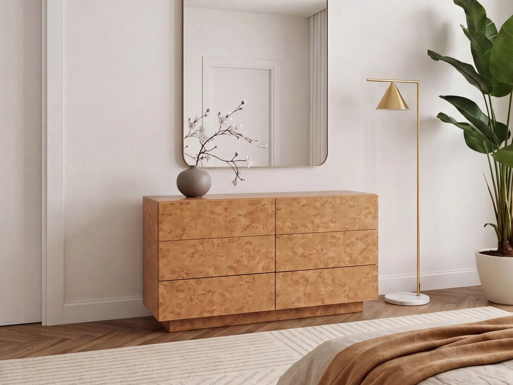 A modern six-drawer wooden dresser with a burl wood grain finish, styled in a minimalist bedroom setting with a matching wall mirror, a gold floor lamp, and a potted plant.