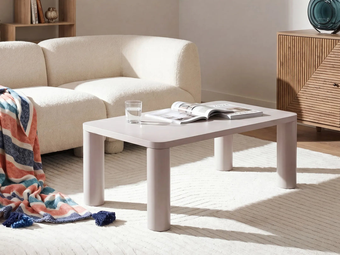 A minimalist rectangular coffee table with a light pink top and matching legs, styled in a bright, modern living room next to a cream boucle sofa.
