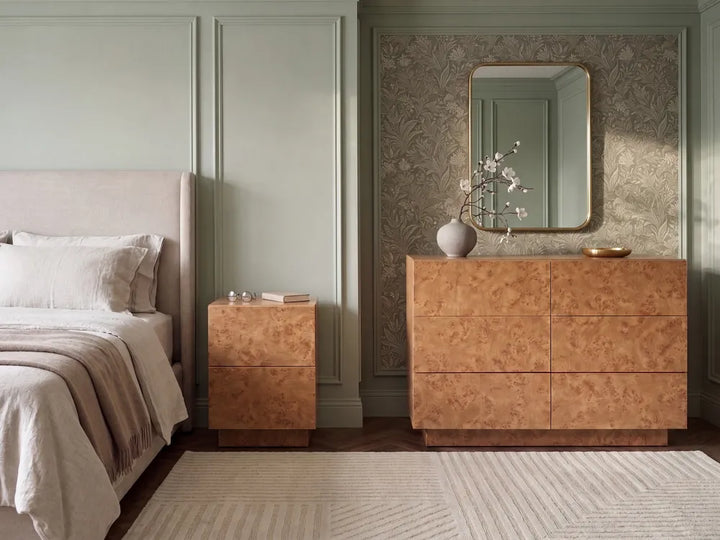 A styled bedroom interior featuring a large copper-brown dresser with a matching wall mirror, a smaller matching nightstand beside a bed, and neutral bedding in a softly lit room with paneled walls and patterned wallpaper.