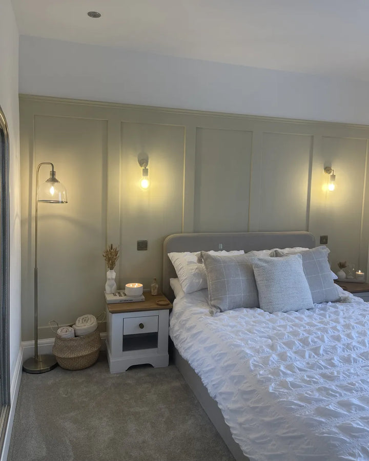 A styled bedroom interior featuring a bed dressed in white textured bedding and light gray pillows, set against a paneled wall with two lit wall sconces. A white nightstand, floor lamp, and woven baskets are visible on the left.