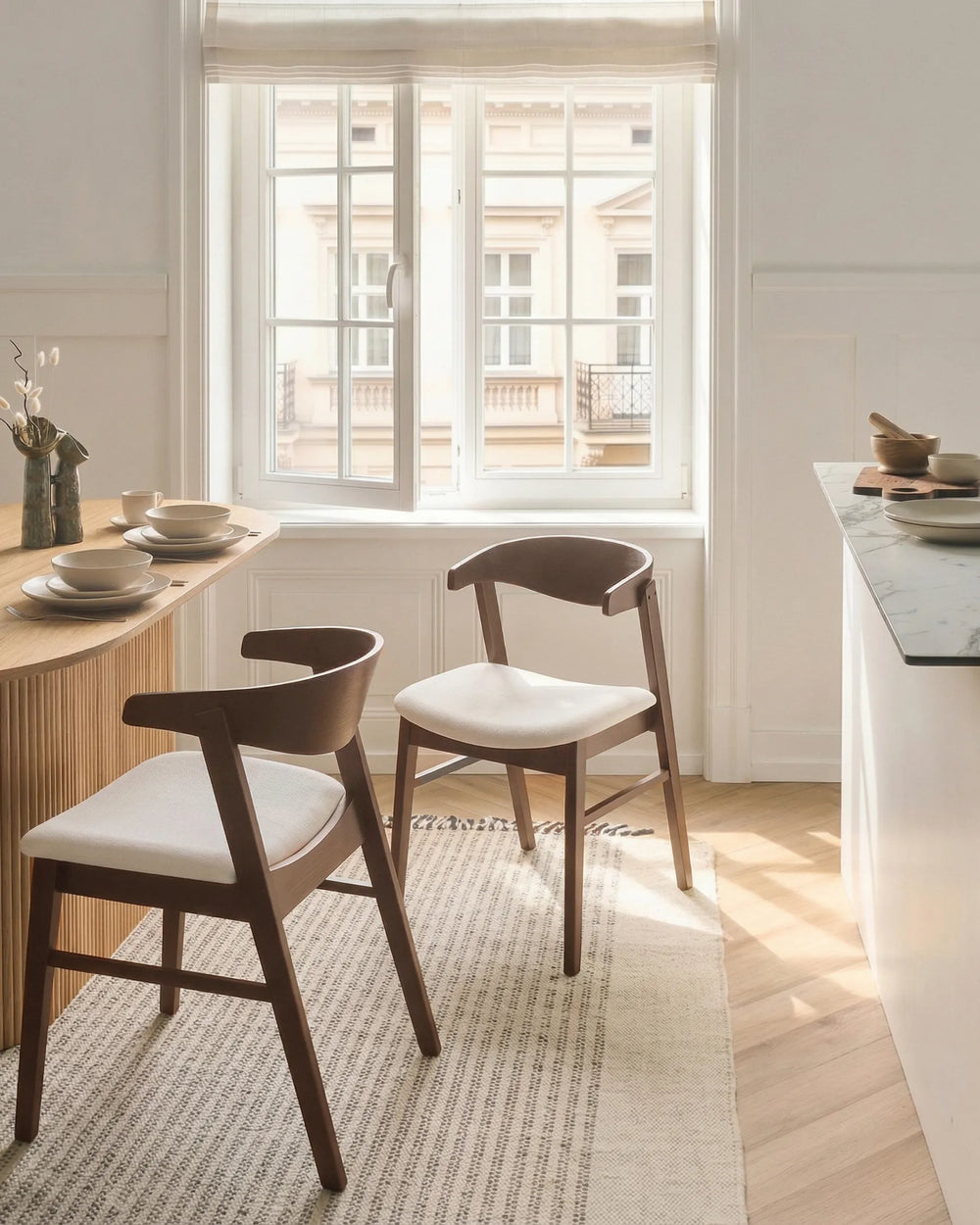 Two modern wooden dining chairs with curved backs and off-white upholstered seats, placed on a light rug in a bright, sunlit dining room near a window.