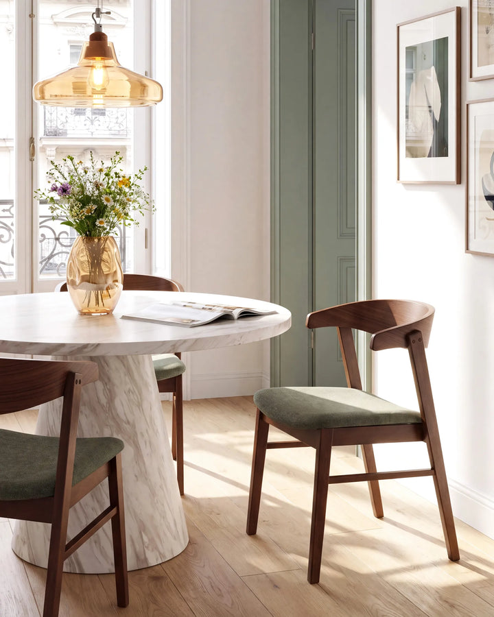 A round white marble dining table with a natural wood base, surrounded by mid-century modern wooden chairs with sage green upholstered seats. A warm amber glass pendant light hangs above, and a vase of wildflowers sits on the table.