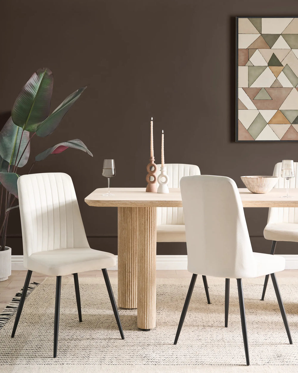 A modern dining room scene featuring a light wood dining table surrounded by four white upholstered dining chairs with black legs, set against a dark brown wall with a framed geometric art print and a potted plant.