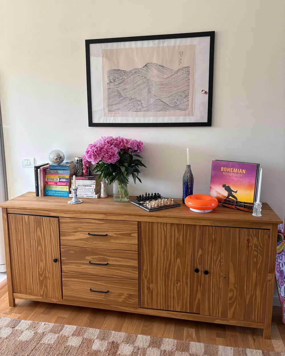 A wooden sideboard or credenza in a warm medium brown finish, styled with books, a vase of pink flowers, a blue candle holder, a chess board, and a Pokémon TCG deck box, set against a cream wall with a framed print above.