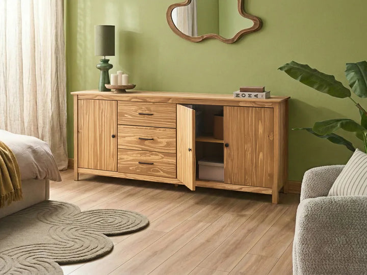 A light natural wood sideboard cabinet with three drawers and two cabinet doors, one of which is open to show shelving, placed in a styled room with a green wall, wood floor, a lamp, and a plant.