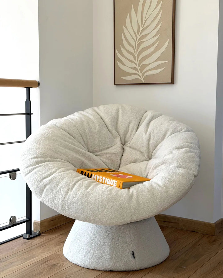 A round, tufted lounge chair upholstered in a soft cream bouclé fabric, set in a minimalist room with a wooden floor and a framed leaf print on the wall.