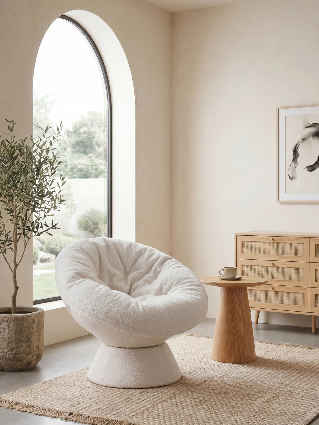 A plush, cream-colored tufted barrel chair with a pedestal base, styled in a minimalist, warm-toned room next to a large arched window.