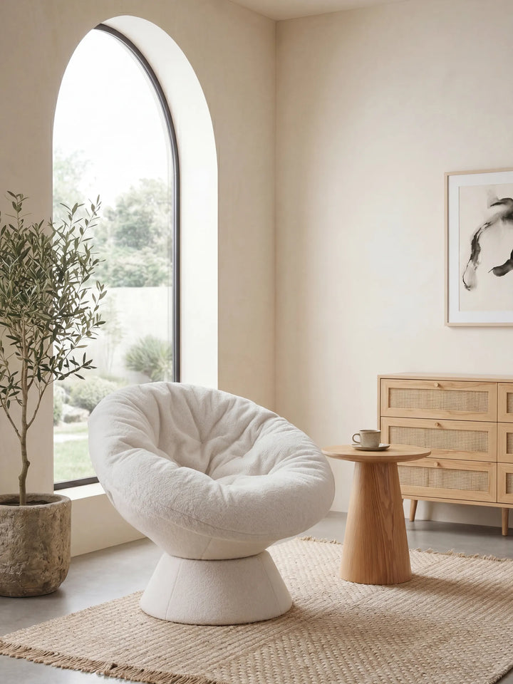 A plush, cream-colored tufted barrel chair with a pedestal base, styled in a minimalist, warm-toned room next to a large arched window.