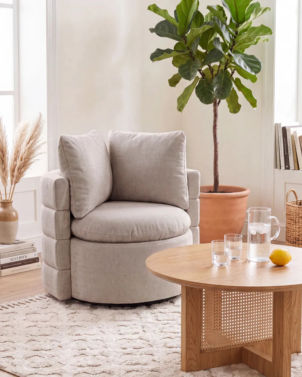 A light beige upholstered accent chair with two matching back cushions, styled in a bright, modern living room next to a round wooden coffee table and a large potted fiddle-leaf fig plant.