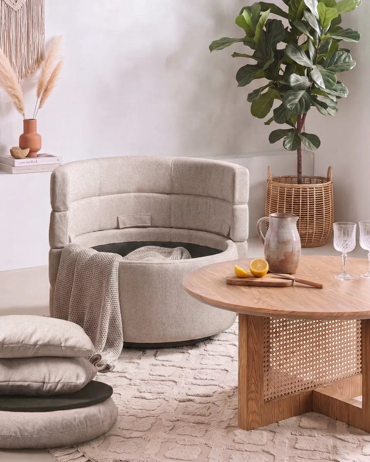 A round upholstered ottoman-style chair in light beige fabric with a tufted back, shown in a styled living room setting next to a wooden coffee table and a potted plant.