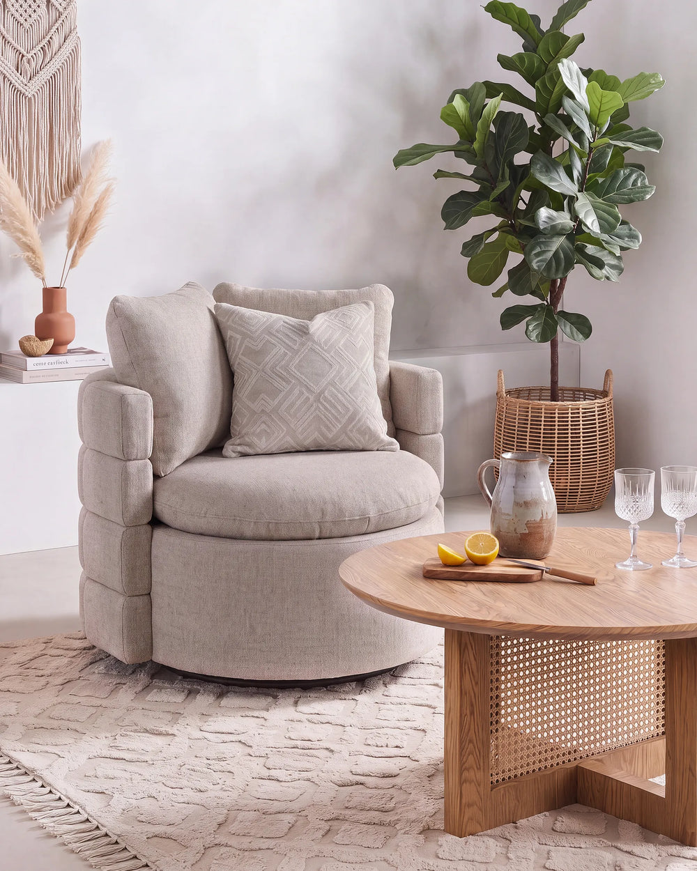 A round upholstered armchair in a light beige fabric with a matching accent pillow, styled in a bright, modern living room next to a wooden coffee table and a potted fiddle-leaf fig plant.