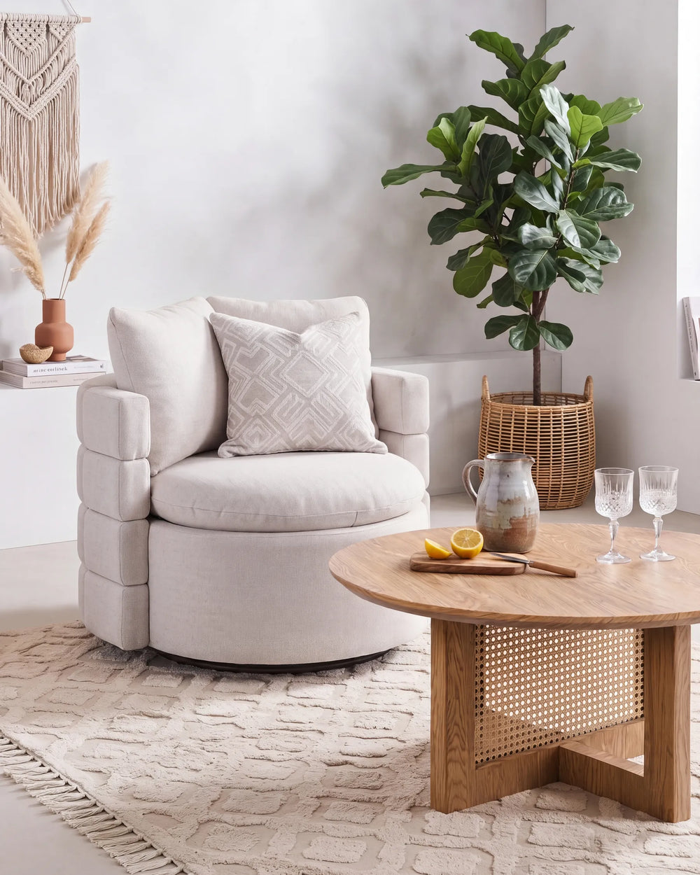 A light beige upholstered accent chair with a geometric patterned throw pillow, styled in a bright, modern living room next to a round wooden coffee table with a woven cane panel base.