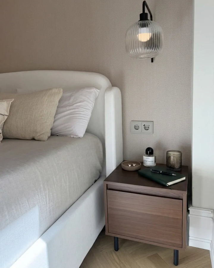 A modern bedroom scene featuring a white upholstered bed with neutral beige and white pillows, a dark wood nightstand with black legs, a ribbed glass wall sconce, and a beige textured wall.