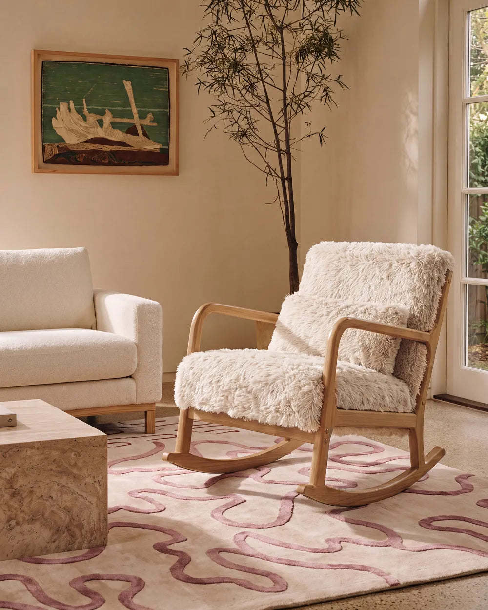 A wooden rocking chair with plush cream sheepskin upholstery, styled in a warm, modern living room next to a white sofa and a patterned rug.