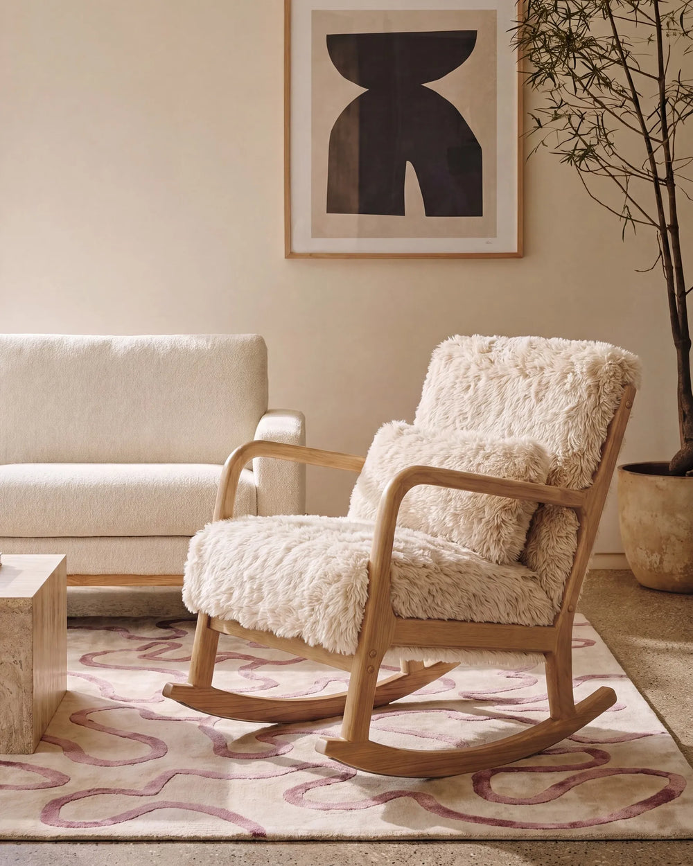 A wooden rocking chair with thick, shaggy cream sheepskin upholstery, styled in a warm, modern living room setting next to a cream sofa and a patterned rug.
