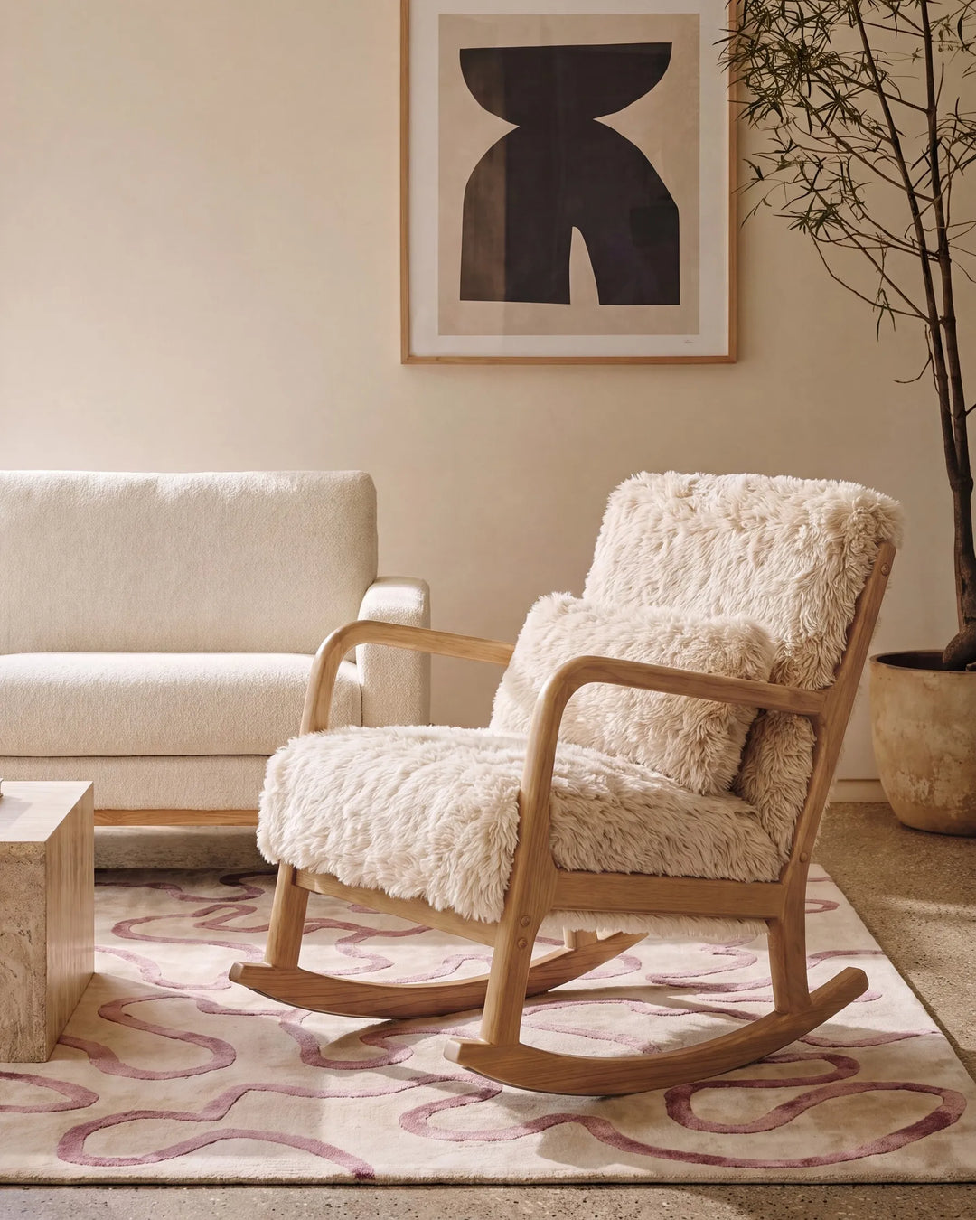 A wooden rocking chair with thick, shaggy cream sheepskin upholstery, styled in a warm, modern living room setting next to a cream sofa and a patterned rug.