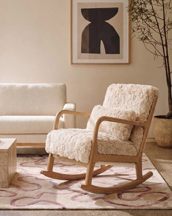 A wooden rocking chair with thick, shaggy cream sheepskin upholstery, styled in a warm, modern living room setting next to a cream sofa and a patterned rug.