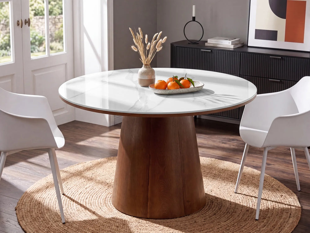 A round dining table with a white marble-look top and a tapered wooden pedestal base, shown in a bright dining room with two white chairs, a woven round rug, and a black sideboard in the background.