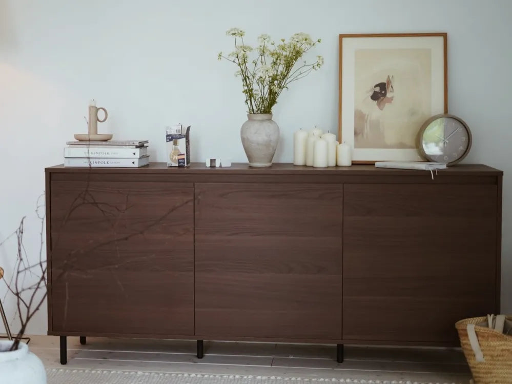 A dark brown wooden sideboard or credenza in a styled room setting, decorated with a vase of flowers, candles, books, and framed art.