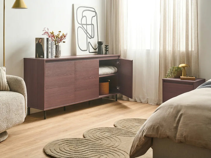 A modern wooden sideboard or credenza in a warm reddish-brown finish, shown in a styled bedroom setting with one door open to reveal storage space. The room includes a bed, armchair, curtains, rug, and bedside table.