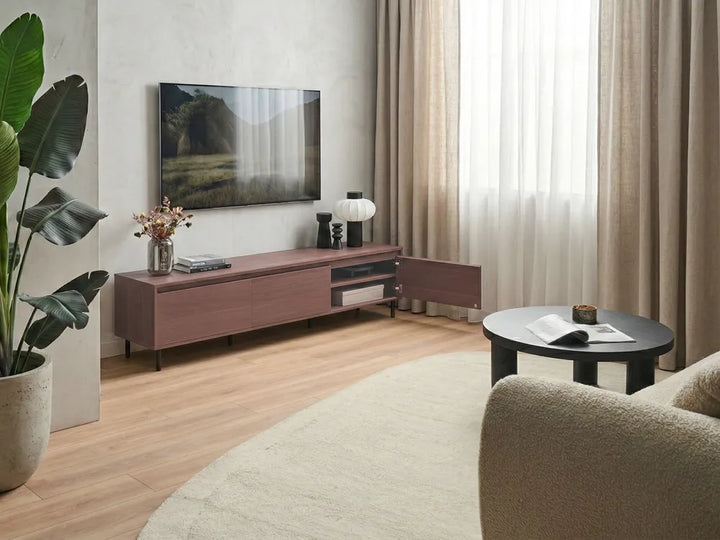 A modern living room scene featuring a low-profile wooden TV console in a dusty rose/brown finish, styled with a wall-mounted flat-screen TV, decorative lamp, and vase. The room has light wood floors, beige curtains, a textured armchair, and a potted plant.