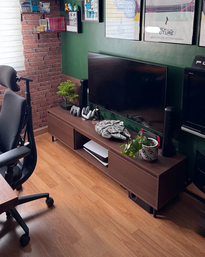 A modern dark brown wooden media console in a home office setting, styled with a large flat-screen TV, gaming console, plants, and accessories against a dark green and brick wall.