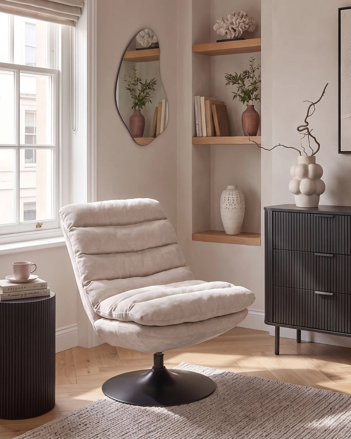 A modern tufted lounge chair in a light beige bouclé fabric with a black metal swivel base, placed in a styled living room setting next to a window and a dark ribbed cabinet.