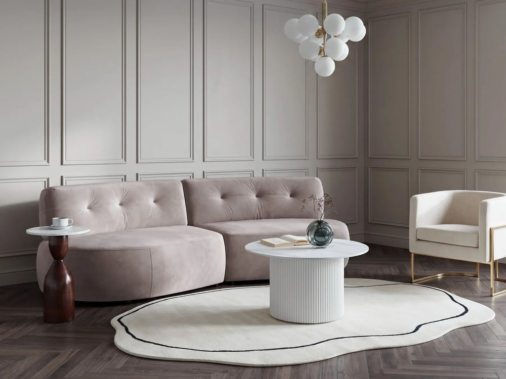 A modern living room featuring a curved, tufted blush pink sofa as the main product, styled with a white ribbed coffee table, a round black-accented rug, a side table, and a white armchair against paneled walls.