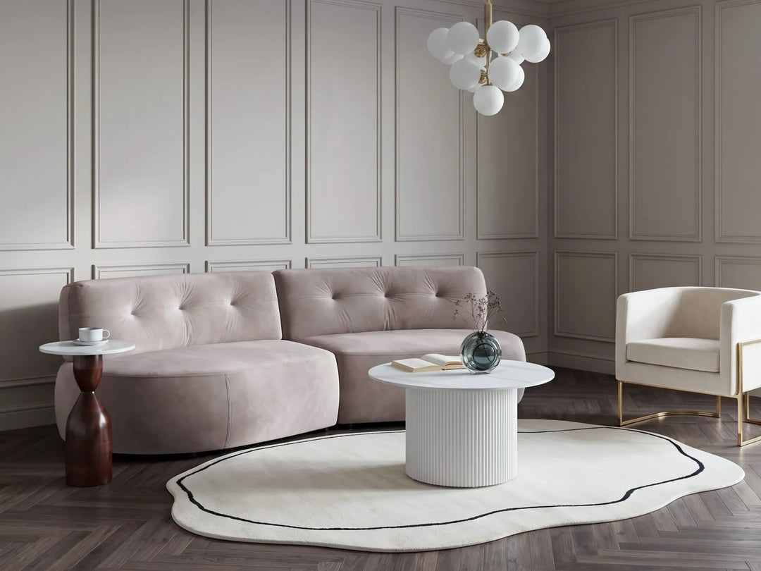 A modern living room featuring a curved, tufted blush pink sofa as the main product, styled with a white ribbed coffee table, a round black-accented rug, a side table, and a white armchair against paneled walls.