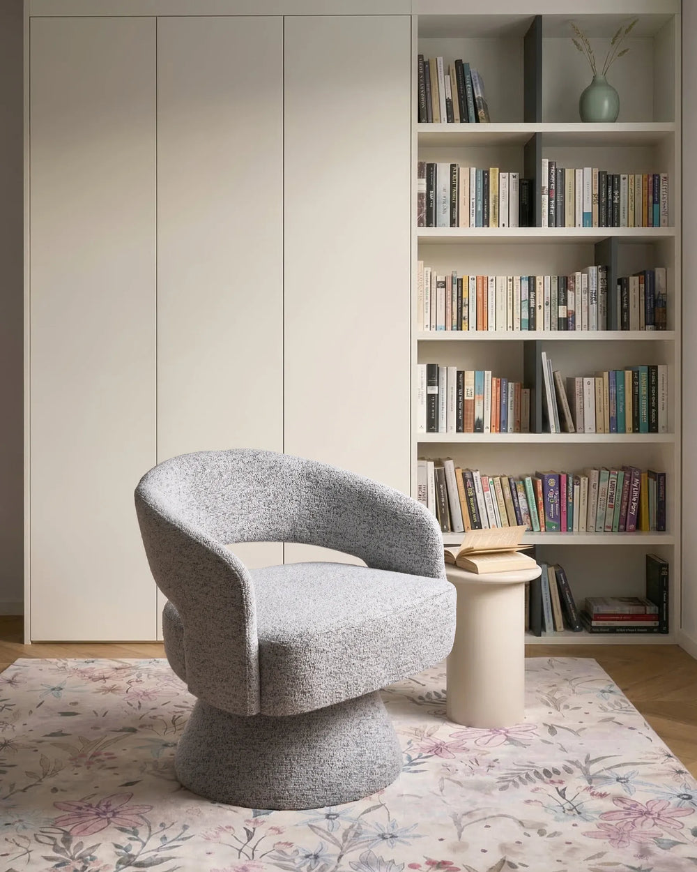 A modern gray bouclé swivel armchair in a styled home office setting with a bookshelf, white side table, and floral rug.