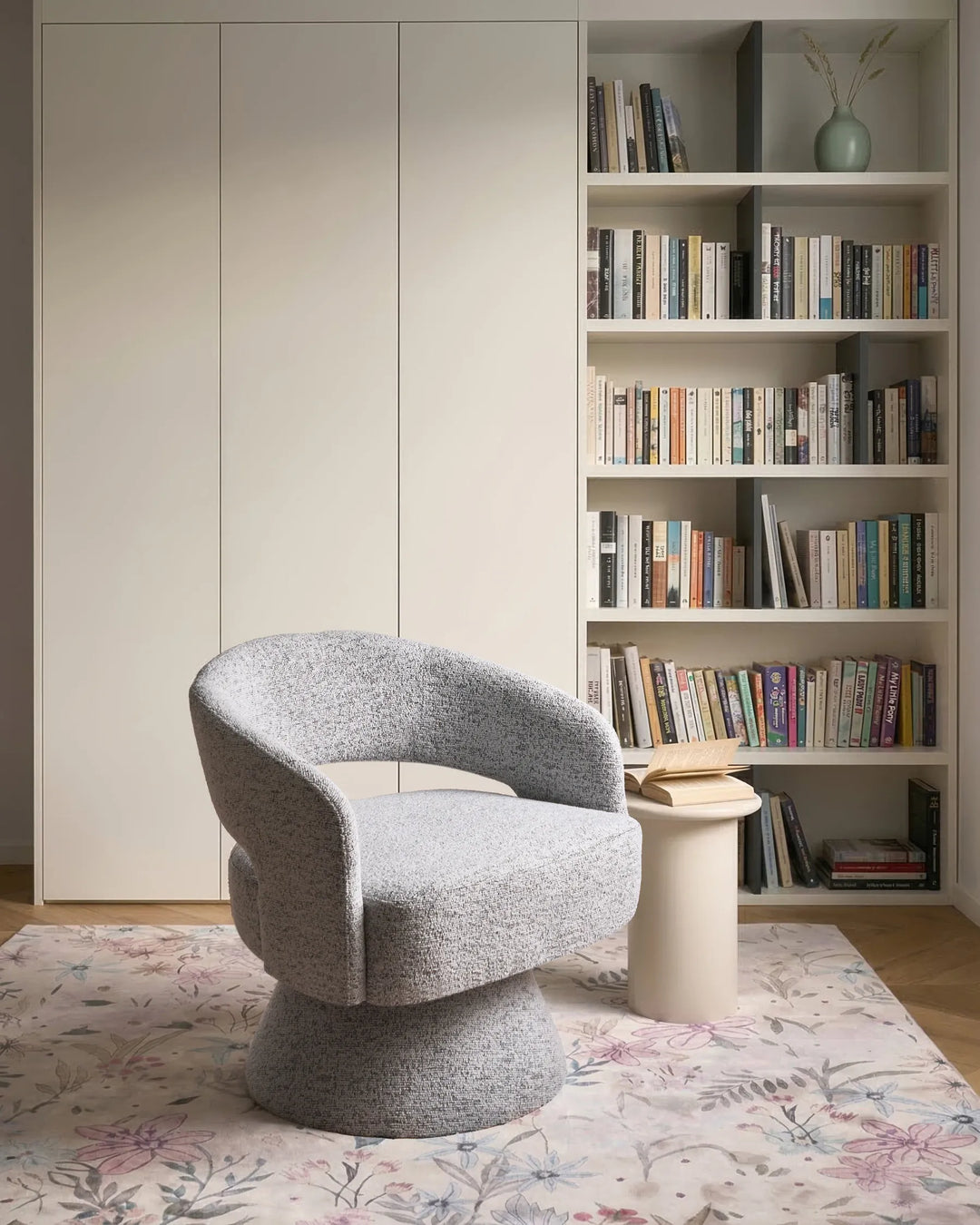 A modern gray bouclé swivel armchair in a styled home office setting with a bookshelf, white side table, and floral rug.