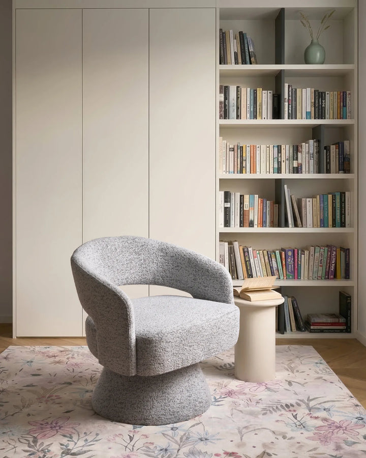 A modern gray bouclé swivel armchair in a styled home office setting with a bookshelf, white side table, and floral rug.
