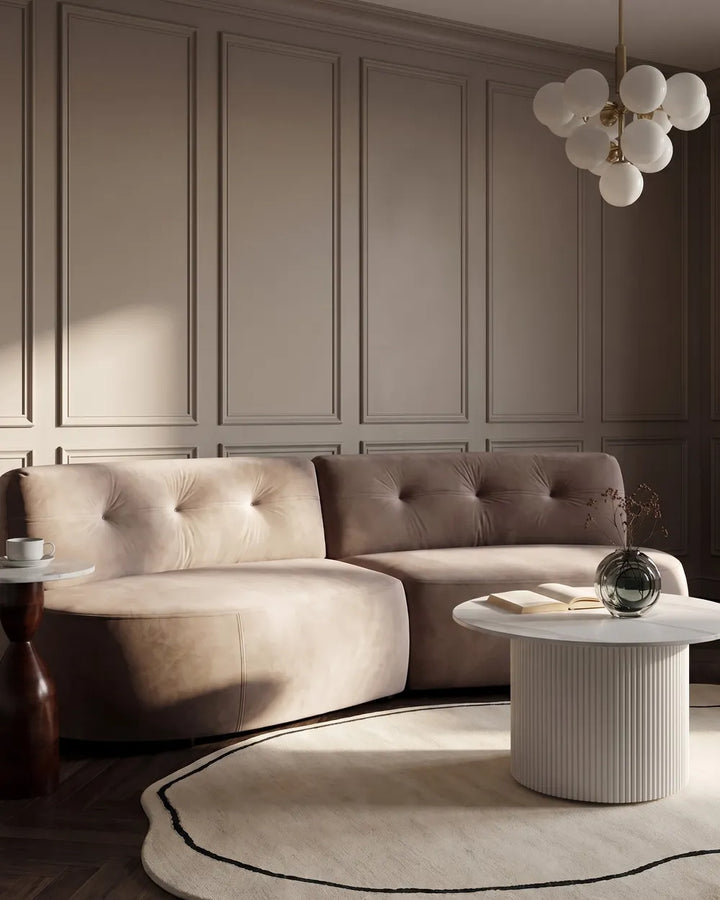 A modern tufted sectional sofa upholstered in a warm taupe-beige velvet, styled in a luxury living room with a white ribbed coffee table, a round area rug, and a decorative wall panel background.