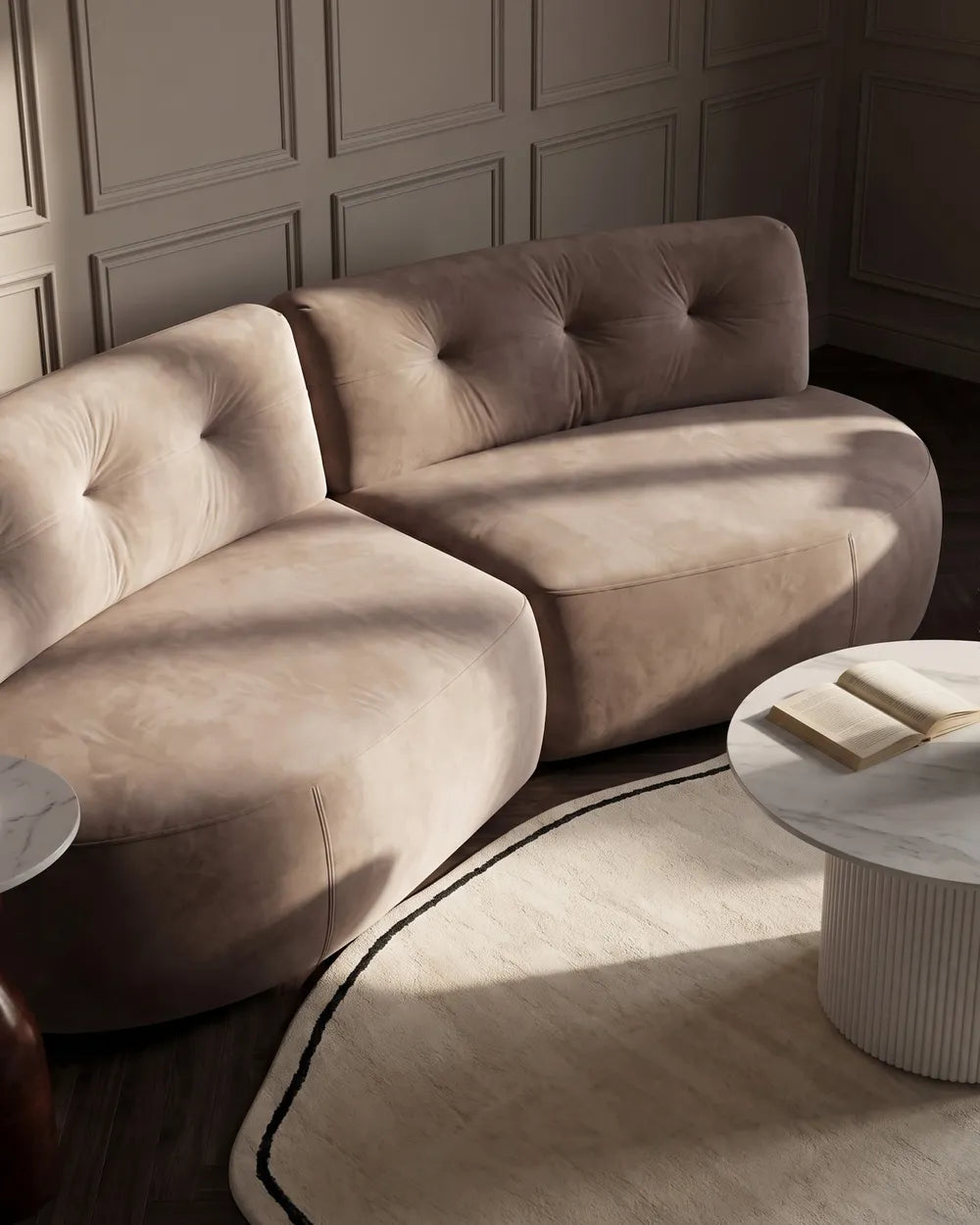 A curved, tufted modular sofa upholstered in a soft taupe velvet fabric, styled in a modern living room setting with a marble coffee table and a patterned rug.