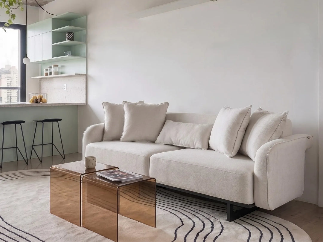 A modern light beige fabric sofa with matching throw pillows in a minimalist living room setting, positioned next to a metallic rose gold coffee table on a patterned rug.
