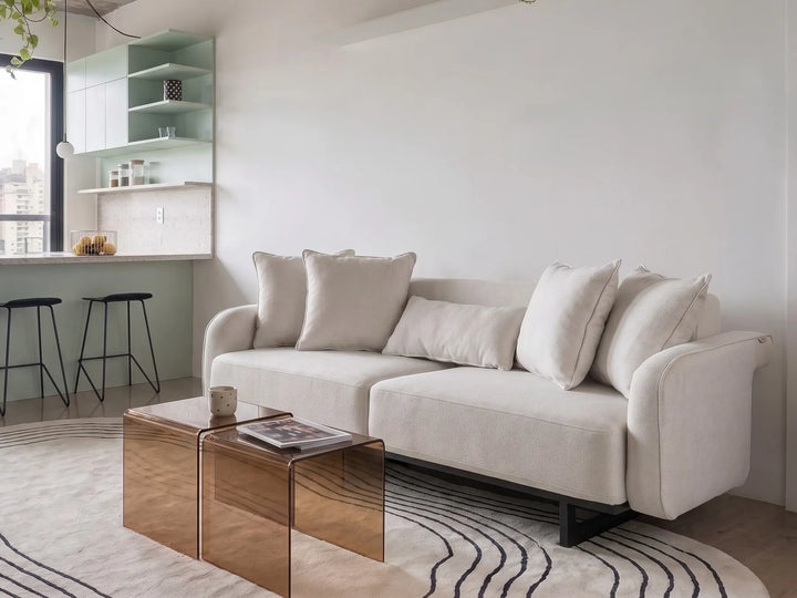 A modern light beige fabric sofa with matching throw pillows in a minimalist living room setting, positioned next to a metallic rose gold coffee table on a patterned rug.