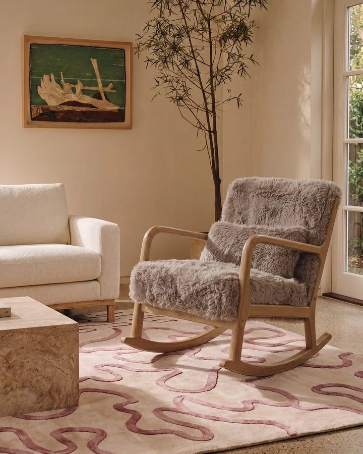 A wooden rocking chair with plush gray faux-fur upholstery in a styled living room setting, next to a white armchair and a patterned rug.