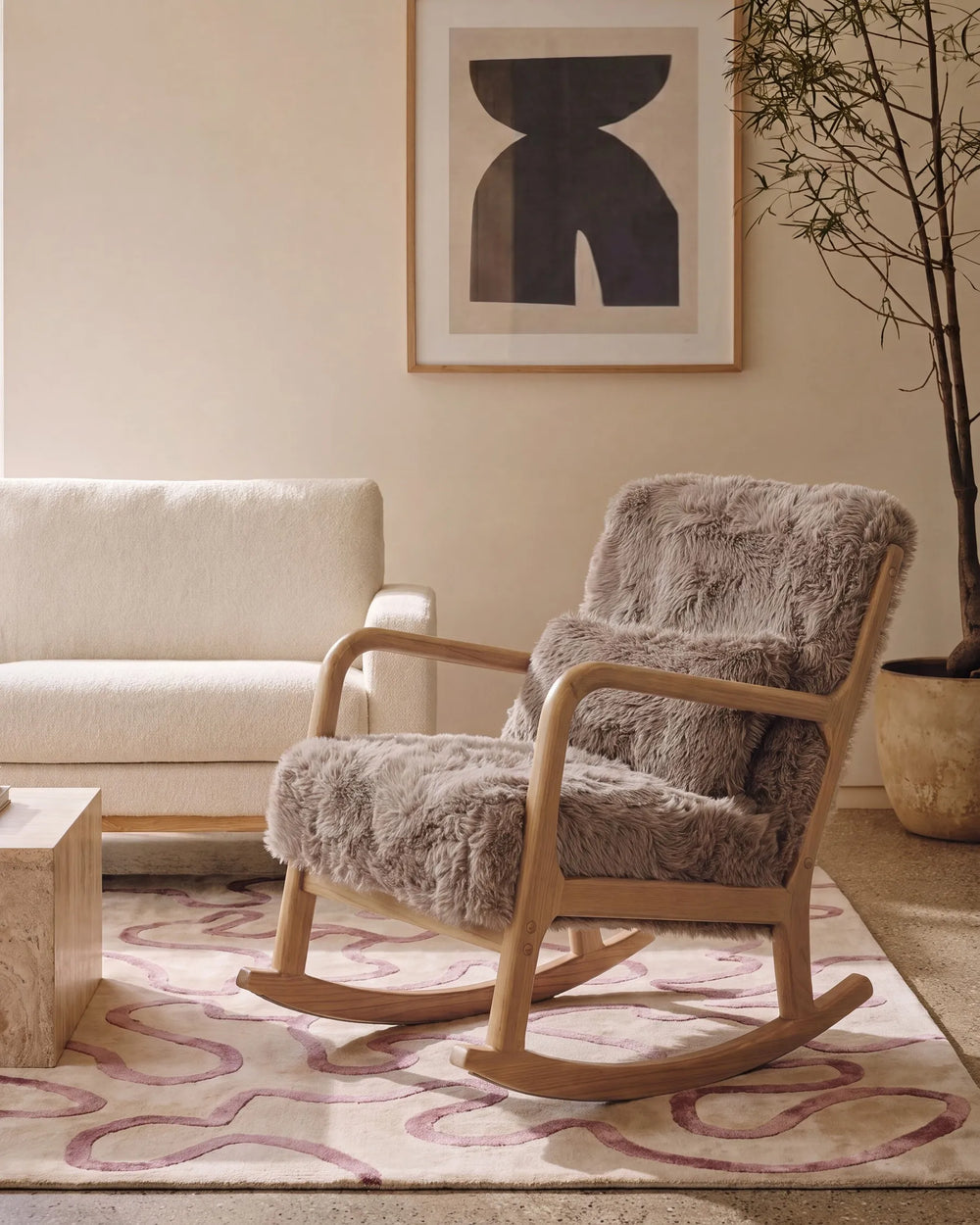 A modern wooden rocking chair upholstered in plush taupe-gray sheepskin, styled in a minimalist living room next to a cream sofa and a patterned rug.