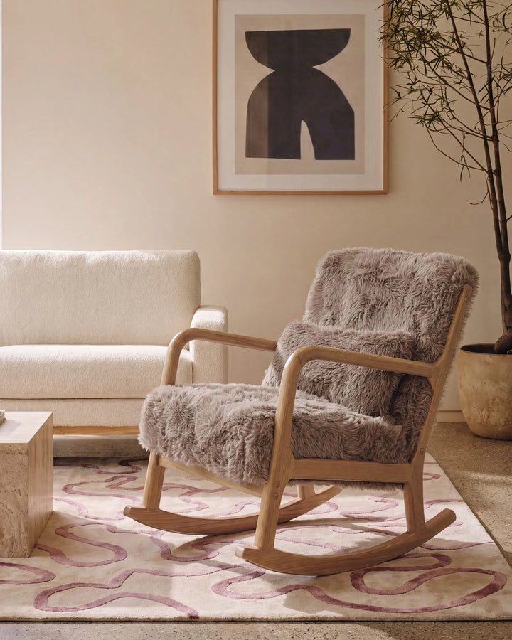 A modern wooden rocking chair upholstered in plush taupe-gray sheepskin, styled in a minimalist living room next to a cream sofa and a patterned rug.