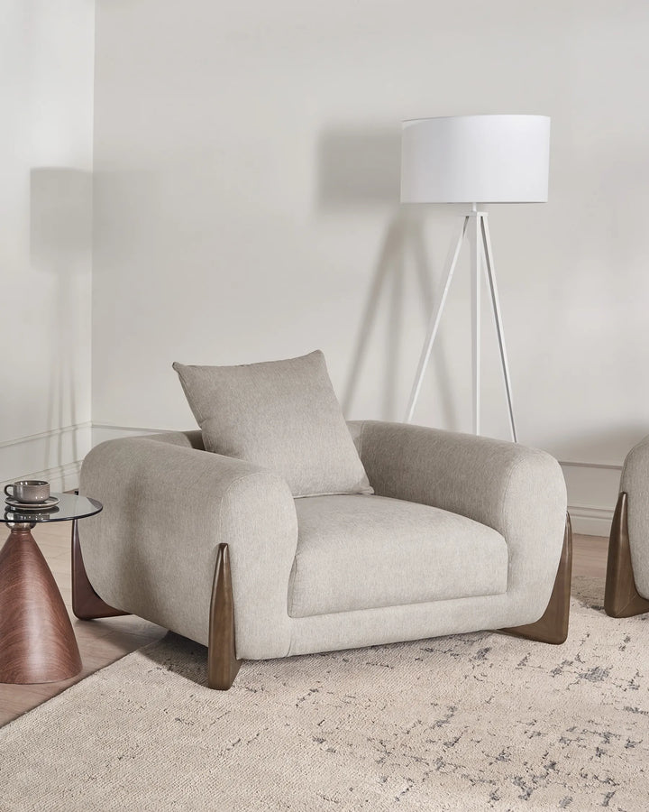 A light beige upholstered armchair with a matching throw pillow, set in a minimalist room with a white tripod floor lamp, a textured rug, and a small side table.