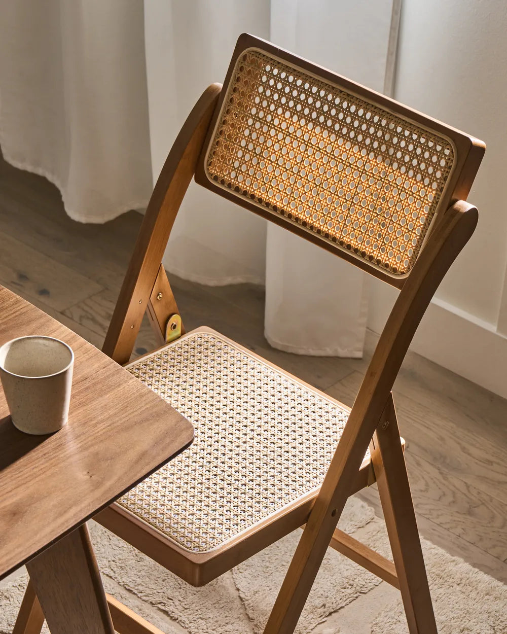 A wooden folding chair with a woven cane backrest and seat, shown in a warm, sunlit room next to a wooden table with a ceramic cup.