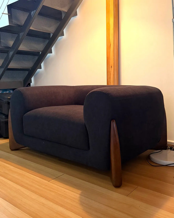 A dark charcoal gray upholstered armchair with curved wooden legs, positioned on a light wood floor in a room with a staircase and warm wall lighting.