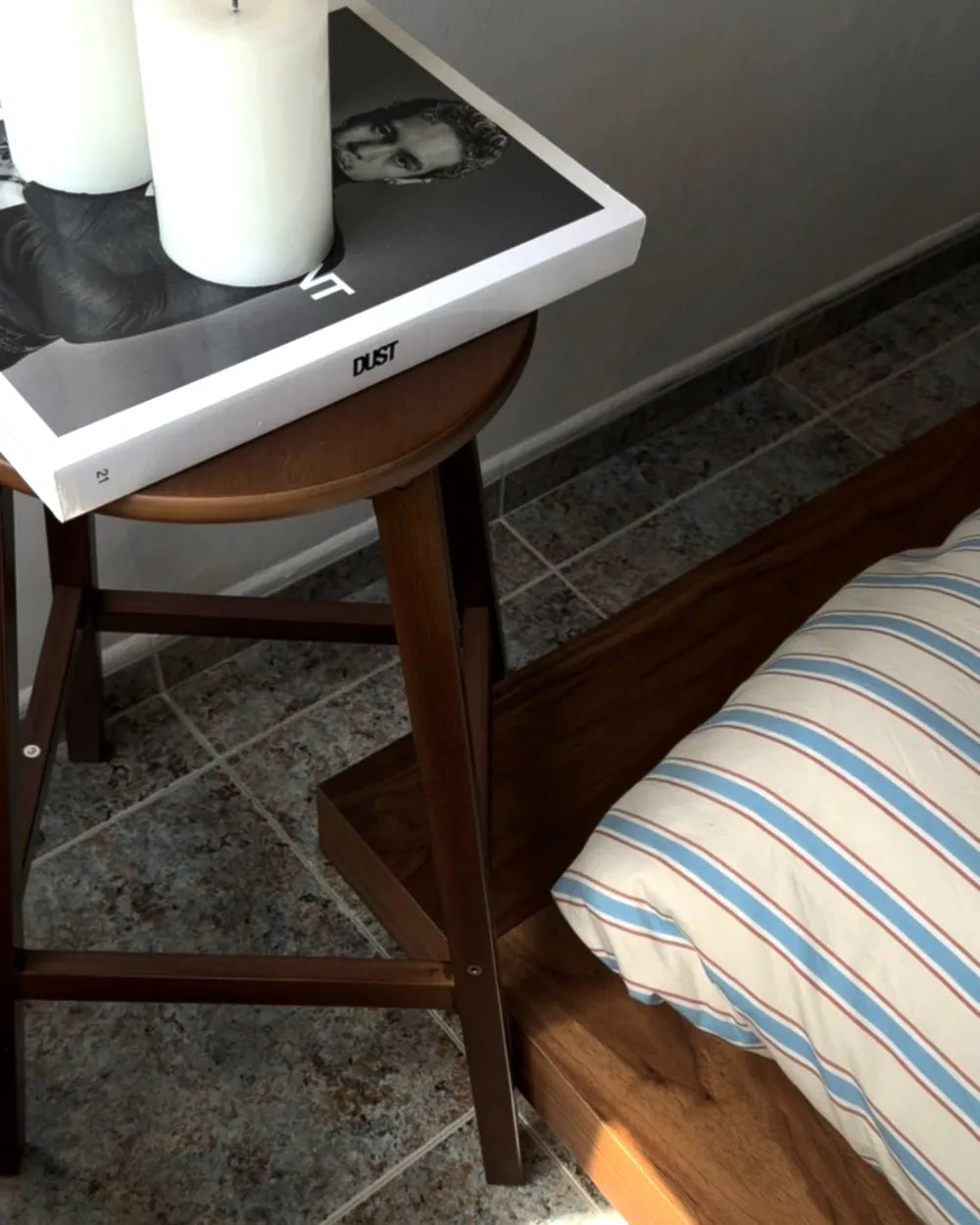 A dark brown wooden side table with a round top and open lower shelf, styled with a white book and two white pillar candles. The table is positioned next to a bed with a blue and white striped pillow, set on a gray tiled floor against a gray wall.