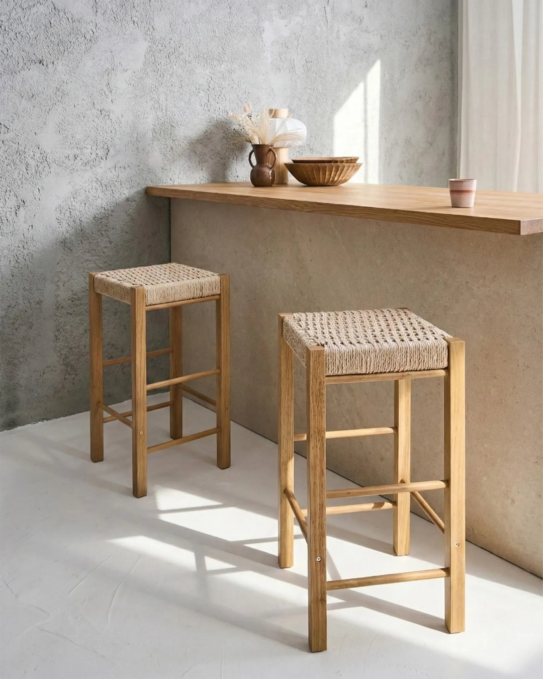 Two wooden bar stools with woven rope seats, positioned in front of a minimalist concrete counter and textured wall.