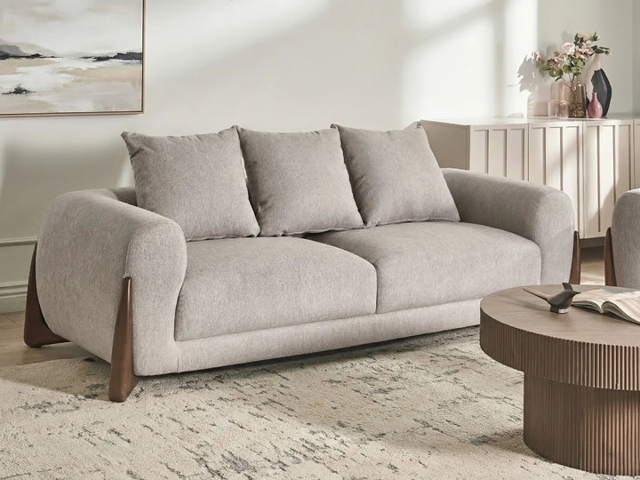 A light gray upholstered loveseat with three matching back cushions, dark wood legs, and rounded arms, shown in a styled living room setting with a round coffee table and neutral wall decor.