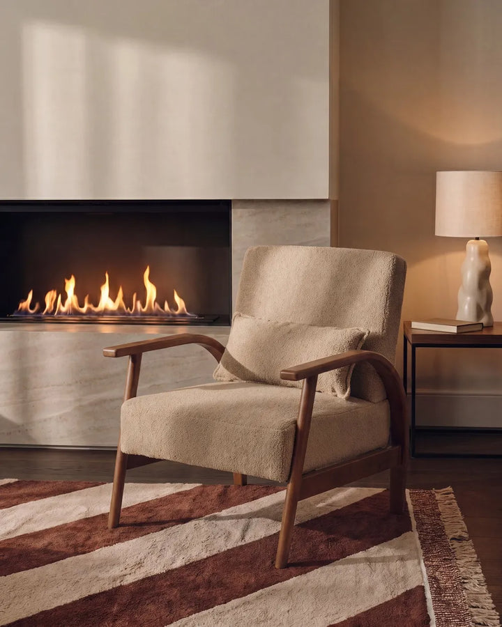 A mid-century modern upholstered armchair with a beige textured fabric and curved wooden arms, styled in a cozy living room next to a fireplace and a lit side table lamp.